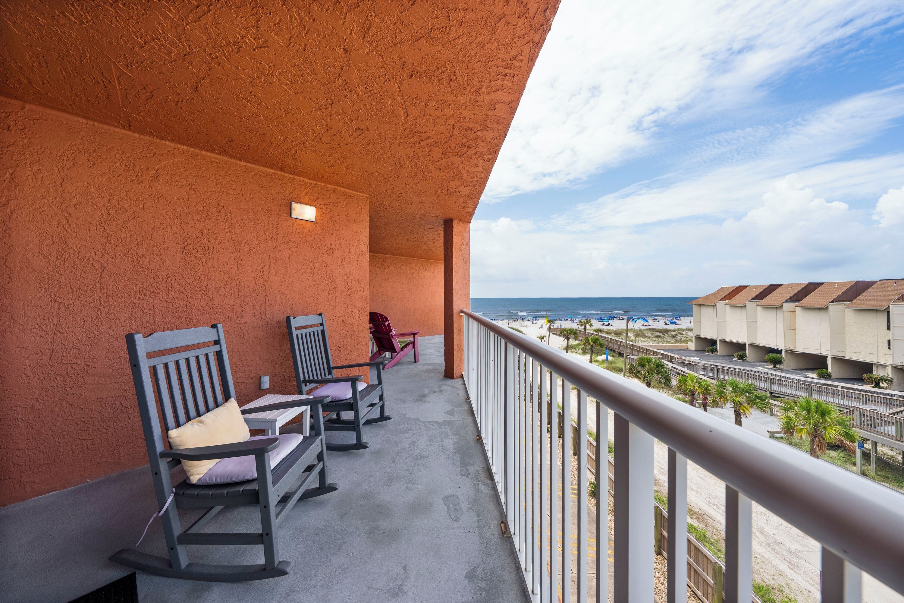 A balcony with rocking chairs and a view of the beach and hotel buildings.