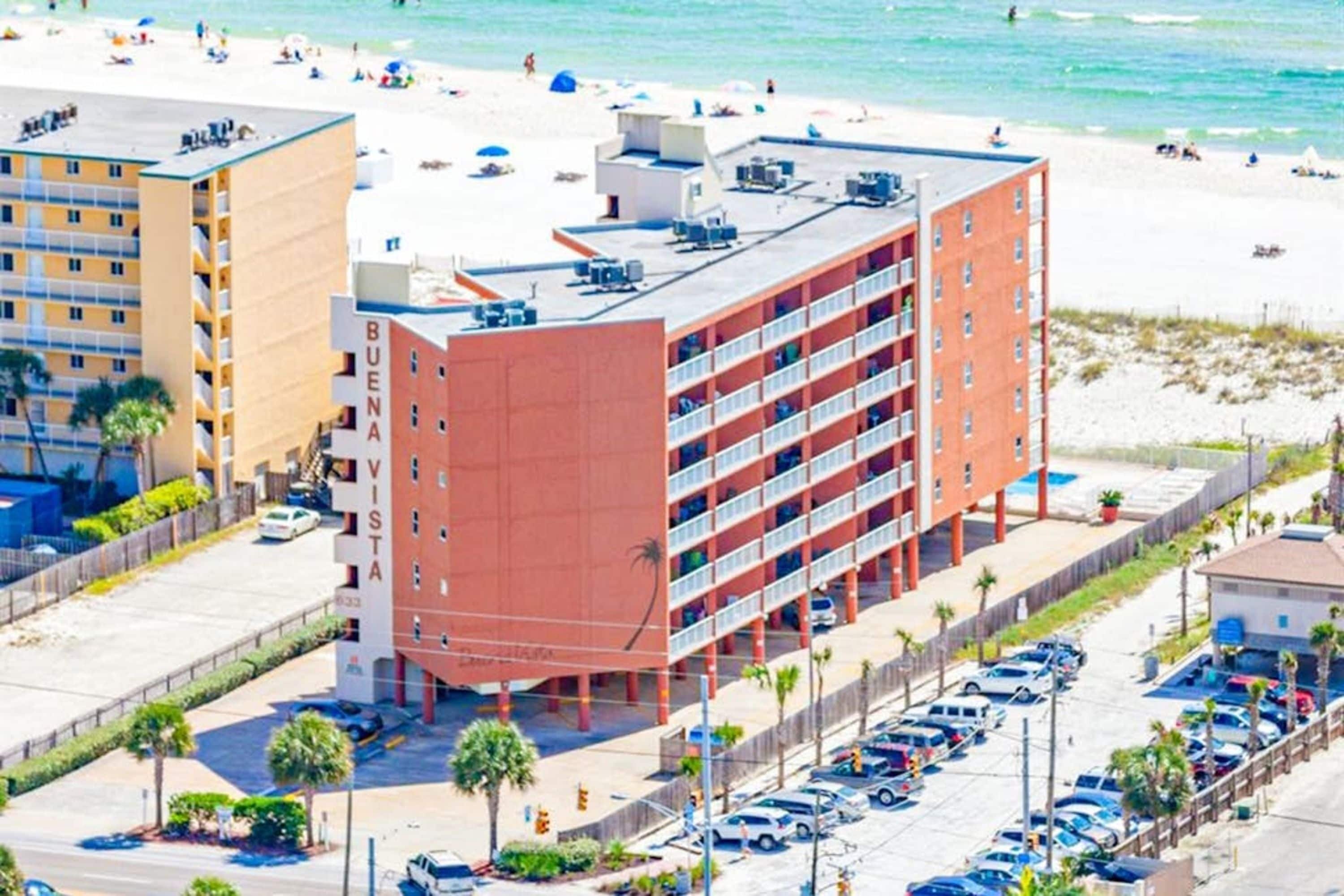 Un hotel de varios pisos con fachada roja, un estacionamiento y una playa al fondo.