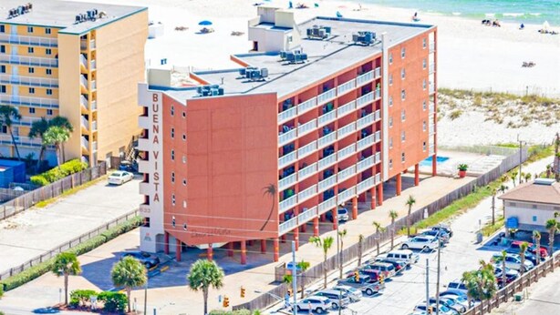 A multi-story hotel with a red facade, a parking lot, and a beach in the background. - Buena Vista 304 (Gulf Shores)