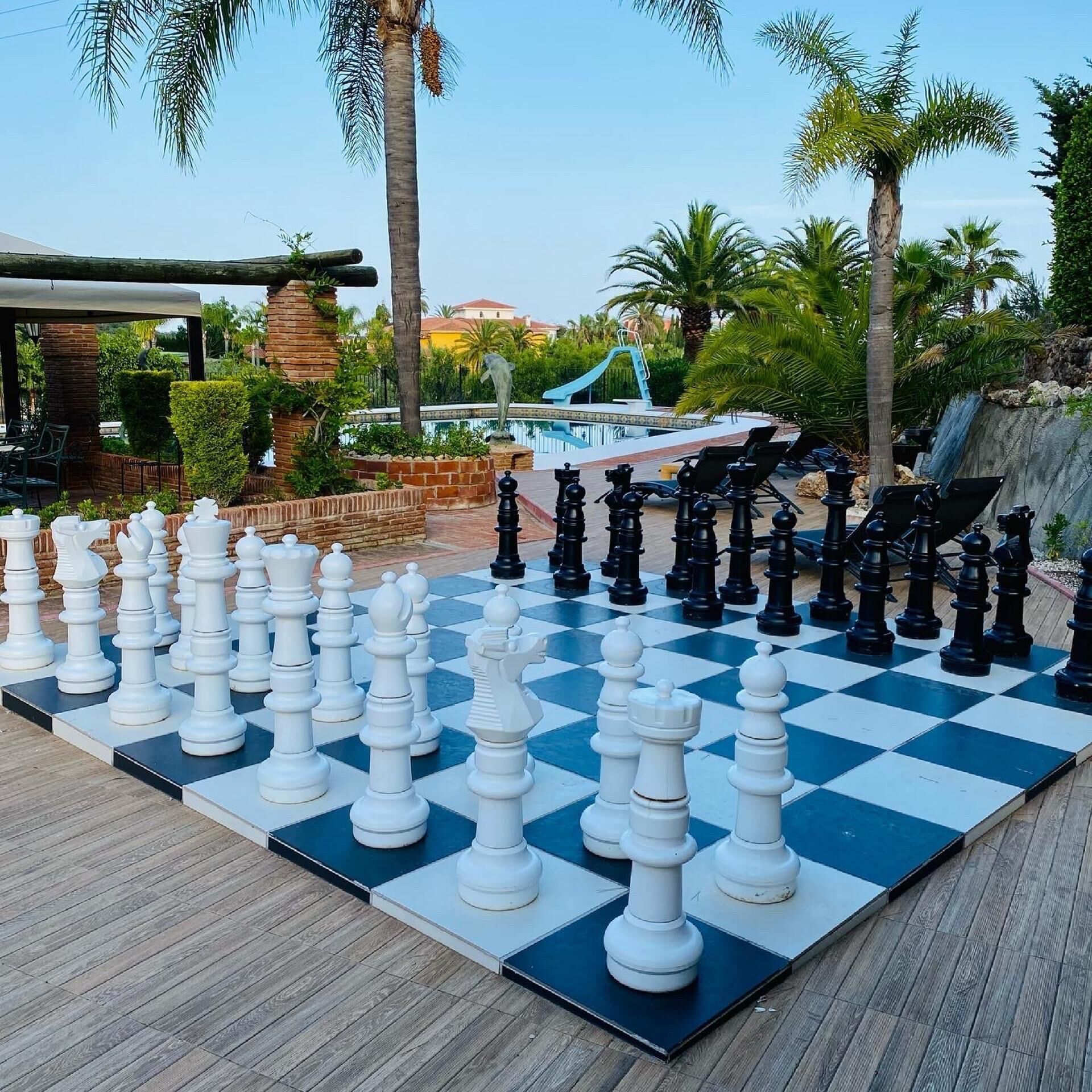 Un tablero de ajedrez grande al aire libre dispuesto sobre una terraza de madera, con una piscina y palmeras al fondo.