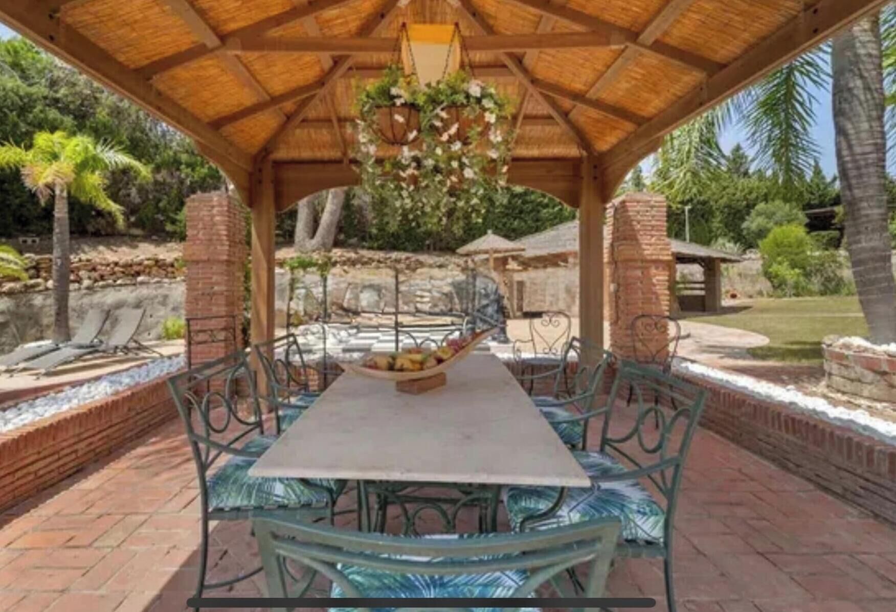 A covered patio with a table and chairs, surrounded by greenery and a brick wall.