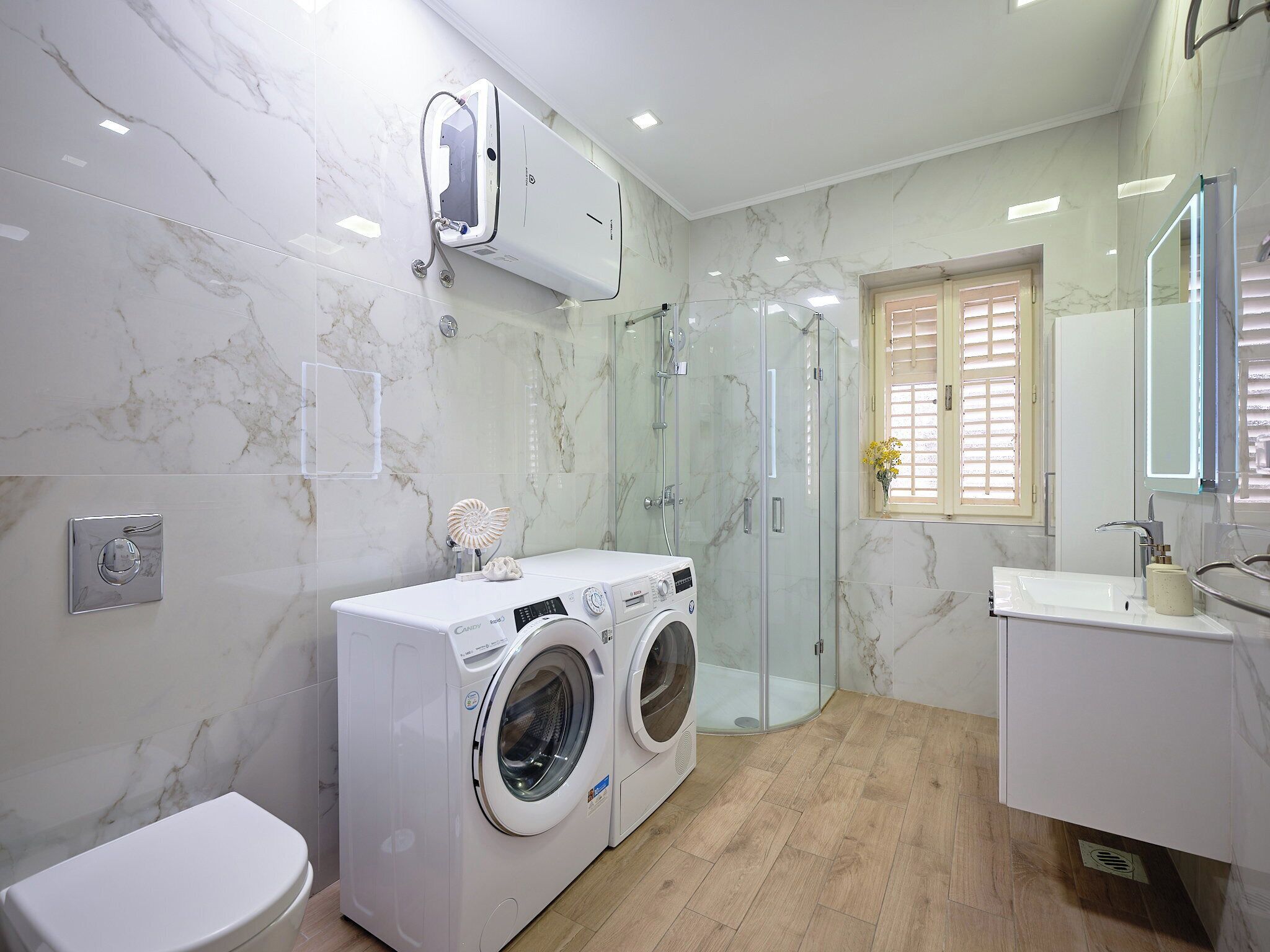 A modern bathroom with a marble wall, a glass shower enclosure, a built-in washing machine, and a white sink.