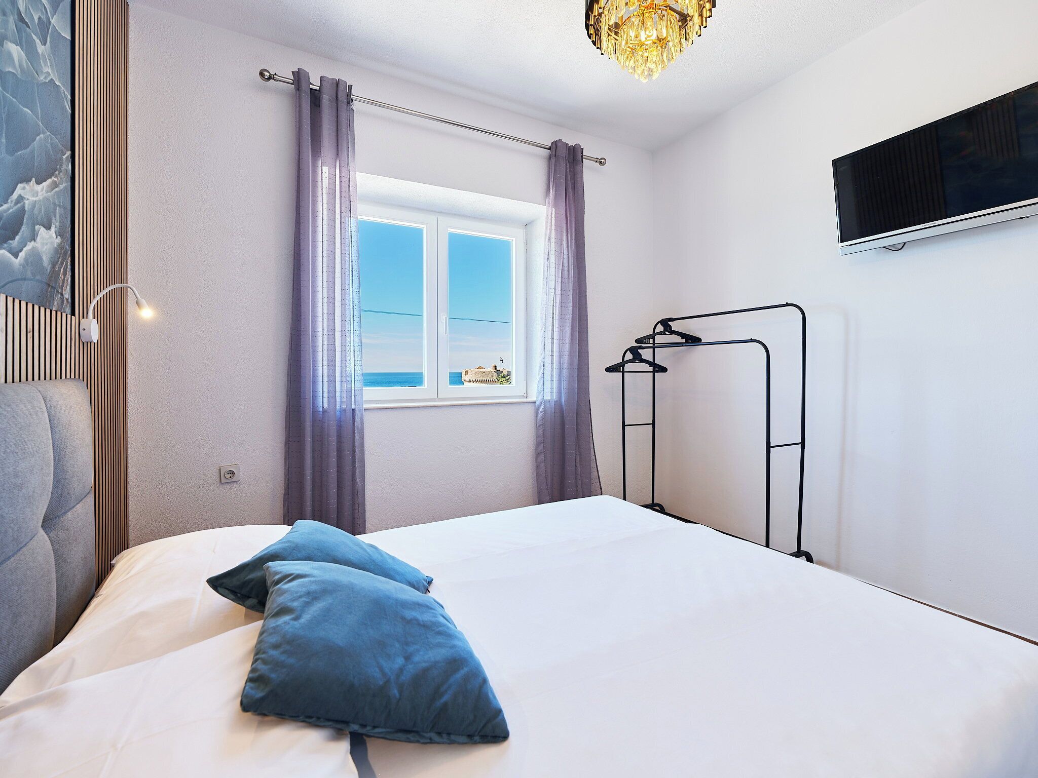 A bedroom with a bed, a window with curtains, a TV mounted on the wall, and a ceiling light fixture.