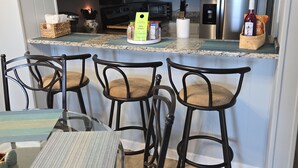 A kitchen with a clock, stainless steel appliances, a dining area with a glass table and chairs, and a basket of snacks. - 2-bedroom Oceanfront condo in peaceful Murrells Inlet with AC comfort (Murrells Inlet)