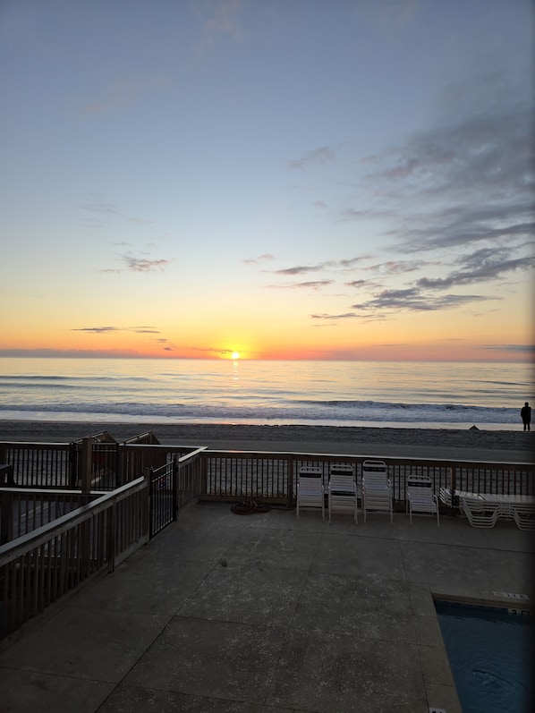 Blick auf einen Strand bei Sonnenuntergang, ein Pool und eine Holzterrasse mit Stühlen.