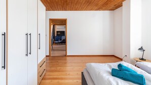A bedroom with a wooden ceiling, a bed with white and blue towels, a desk with a lamp, and a view into another room with a blue chair.
