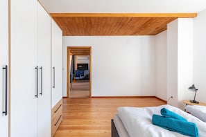 A bedroom with a wooden ceiling, a bed with white and blue towels, a desk with a lamp, and a view into another room with a blue chair.