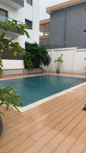 Ein Schwimmbecken mit Holzterrasse, umgeben von Topfpflanzen und einem modernen Gebäude im Hintergrund.
