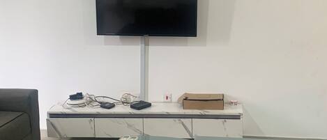 Una sala de estar con un televisor de pantalla plana montado en la pared, una mesa de centro de vidrio con controles remotos, un sofá gris y un tapete estampado.
