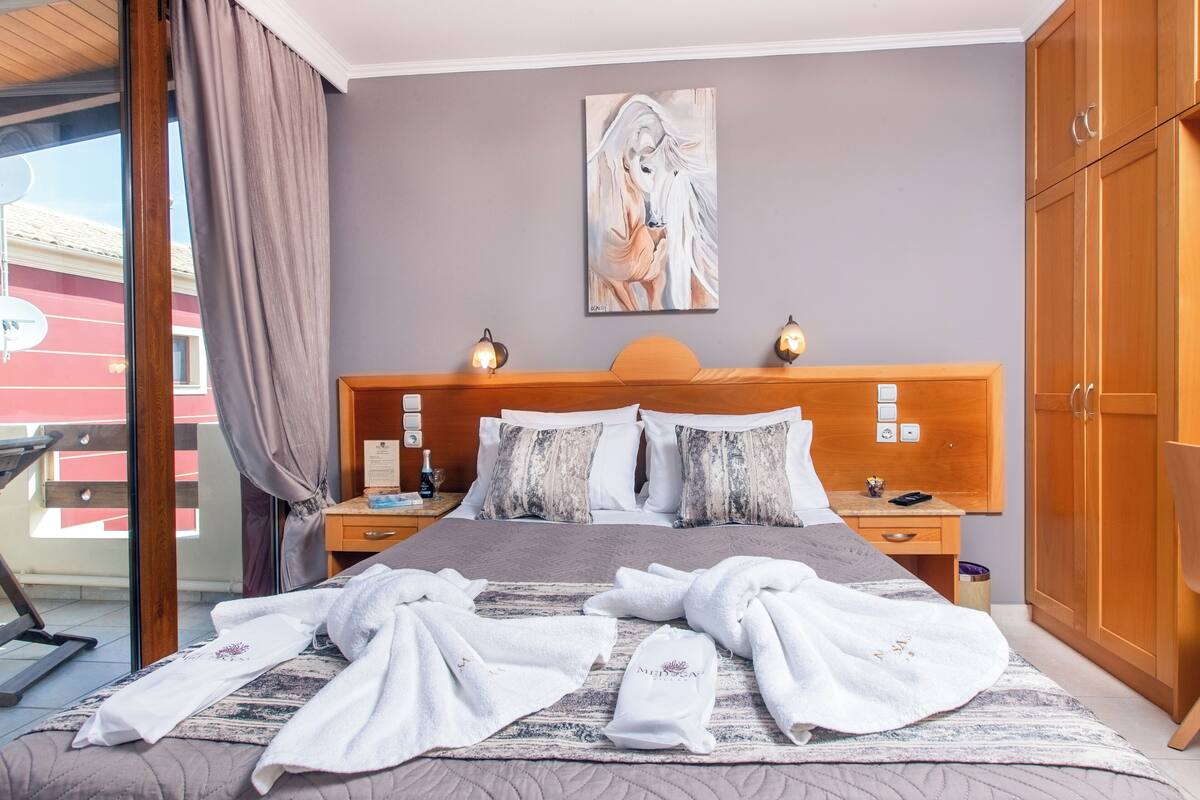 Une chambre d’hôtel avec un lit, un tableau au mur, une tête de lit en bois, deux tables de chevet avec des lampes et une vue à travers une porte vitrée.
