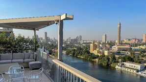 A balcony with outdoor furniture overlooking a river and cityscape. - AB N401 Spacious 2BR Duplex with Partial Nile View (Giza Governorate)