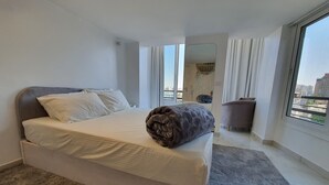 Moderni hotellihuone, jossa on suuri sänky, harmaa matto, harmaa sohva ja ikkunasta avautuva näkymä kaupunkiin.