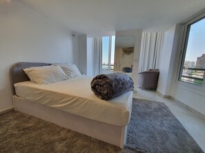 Ein modernes Hotelzimmer mit einem großen Bett, einem grauen Teppich, einer grauen Couch und Blick auf die Stadt durch das Fenster.