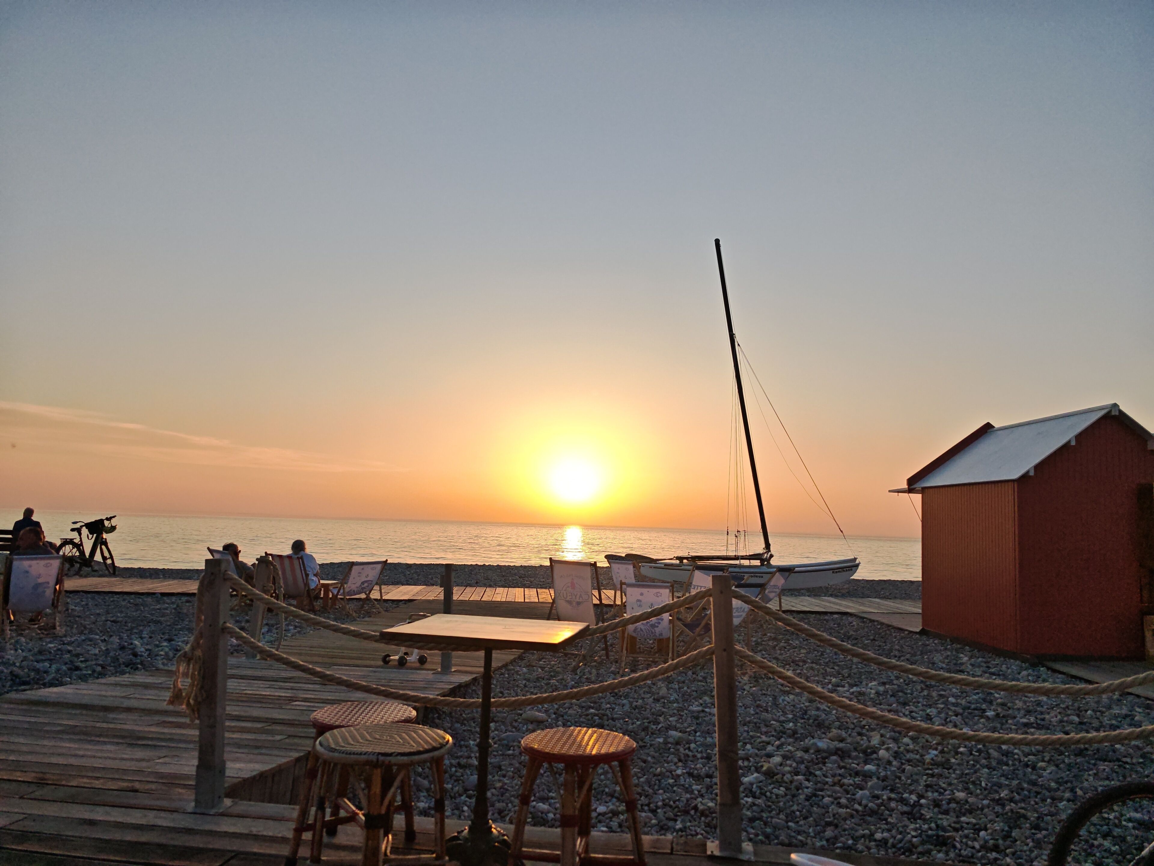 A beachside setting with a wooden deck, wicker chairs, and a small building.