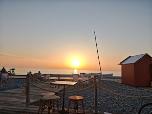 A beachside setting with a wooden deck, wicker chairs, and a small building.