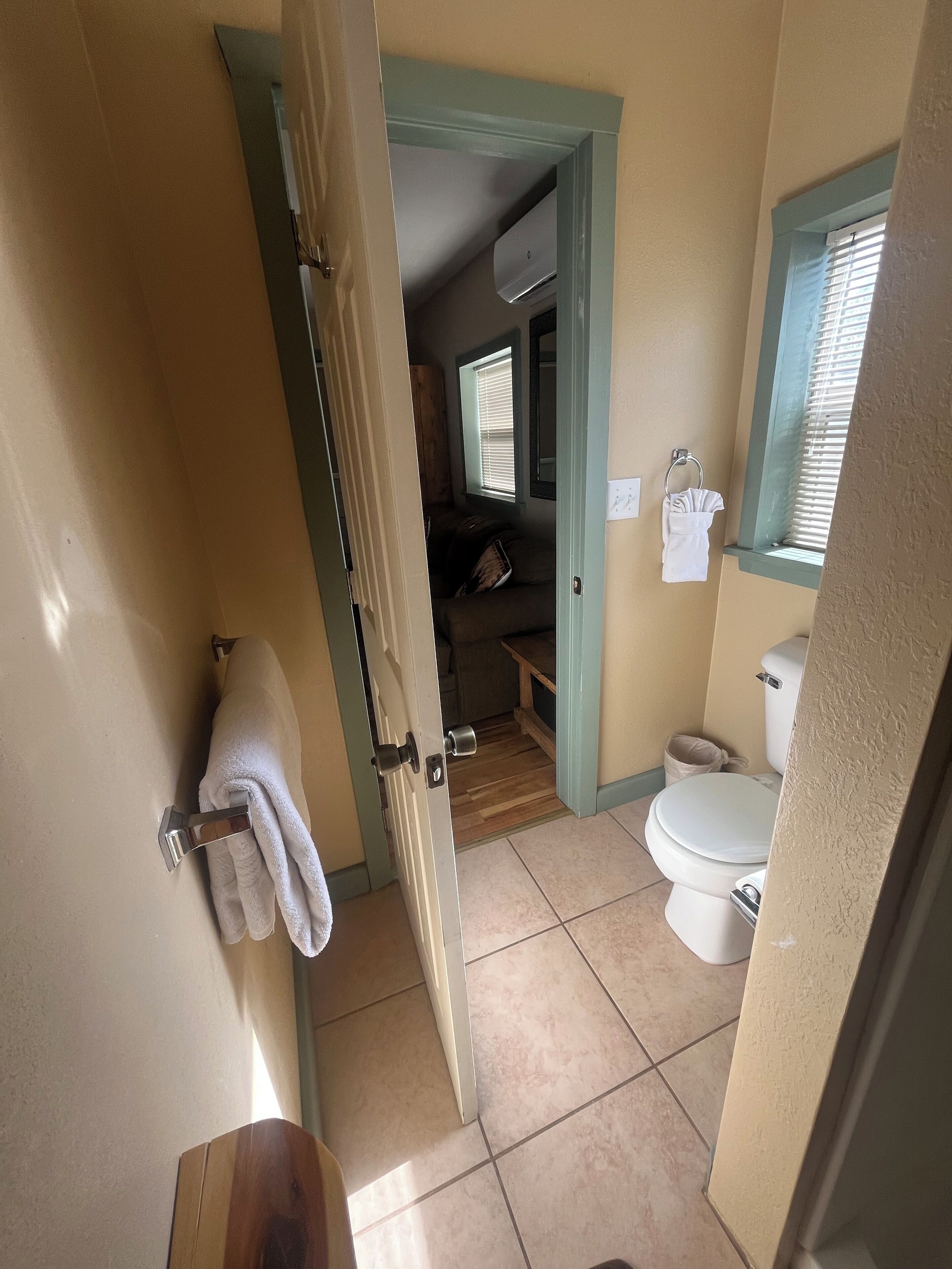 A bathroom with a toilet, a towel rack, a window with blinds, and a door leading to another room.