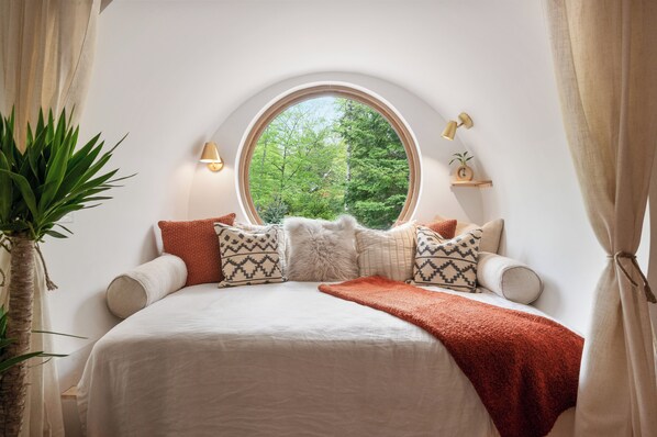 A bedroom with a large round window, a bed with multiple pillows, a red throw blanket, and two wall-mounted lamps. - Creekside Couple's Escape w/Hot tub, Sauna Near Lake (East Jewett)