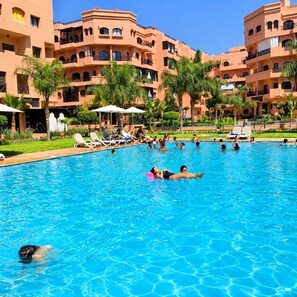 A swimming pool with multiple levels of buildings in the background, surrounded by palm trees and lounge chairs. - Charming 2-bedroom Apartment With Mountain Views in Marrakesh (Marrakech)