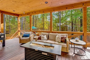 Una terraza de madera con un brasero, mobiliario exterior y vistas a un bosque.