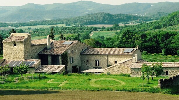 Un bâtiment en pierre doté d’une tour, entouré d’une végétation luxuriante et de champs.