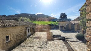 Une cour bordée de bâtiments en pierre, un champ verdoyant et une colline en arrière-plan.