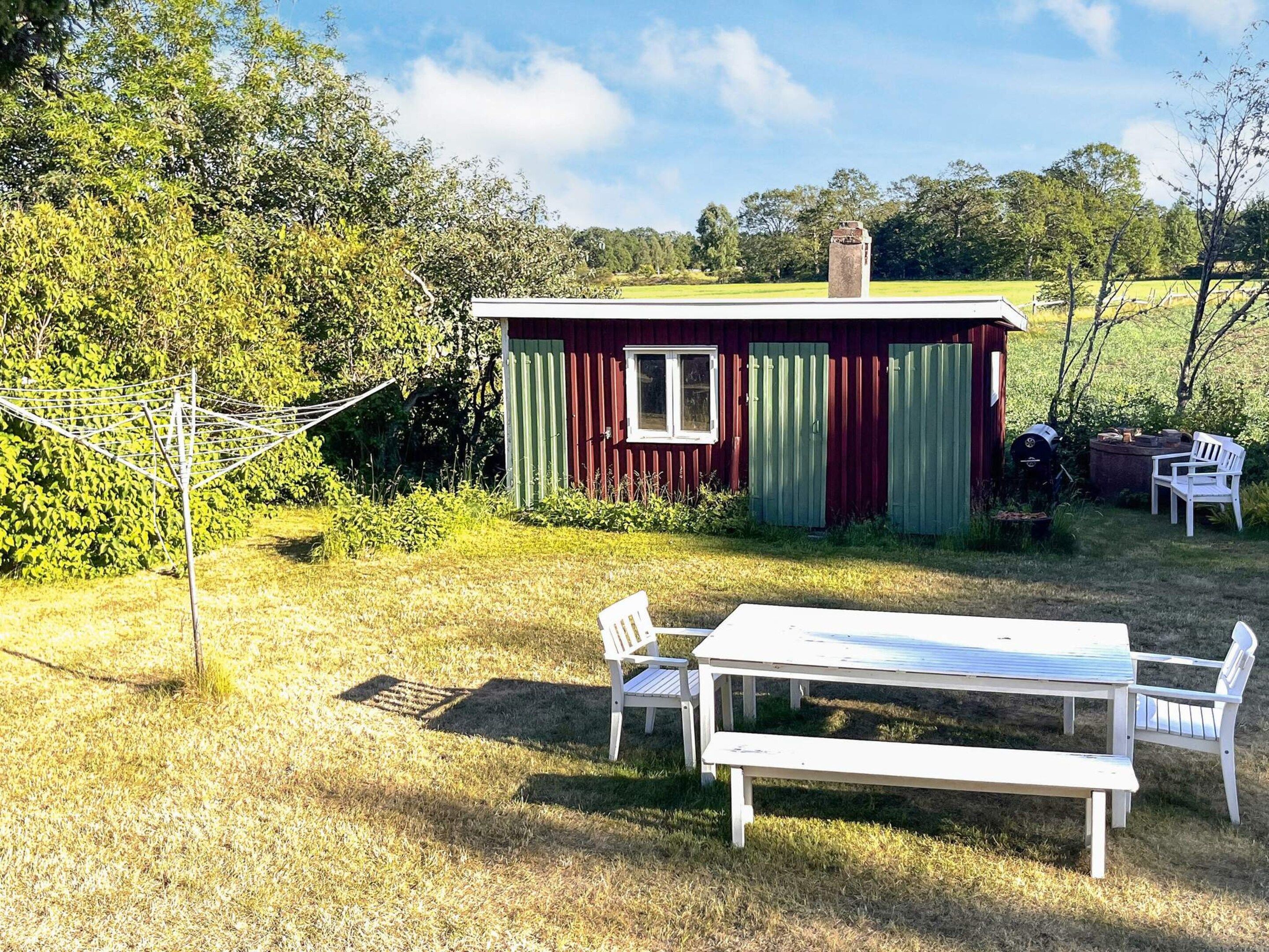Una pequeña casa de madera con fachada roja y verde, una mesa blanca y dos sillas, y un tendedero en una zona con césped.