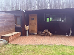 Eine Holzhütte mit einer Holztür, einer Bank, einer Metallvase mit getrockneten Pflanzen und einem Holzstapel.