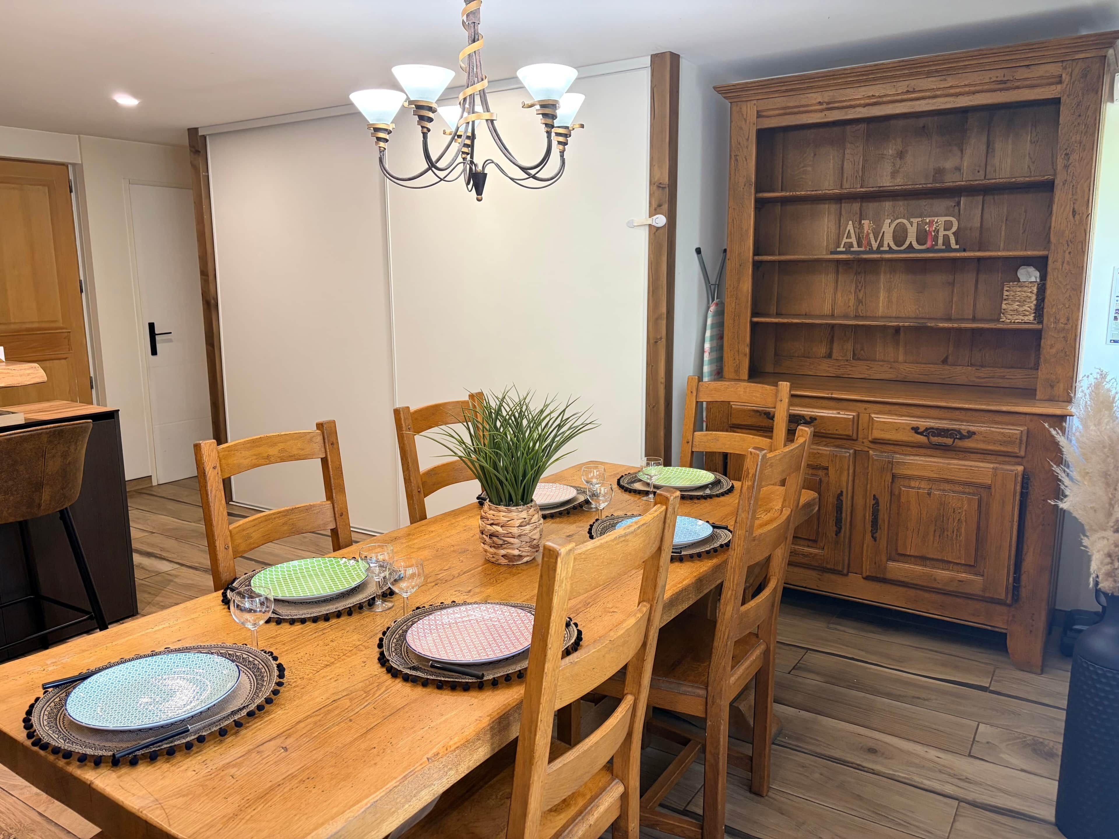 Uma sala de jantar com mesa de madeira posta para quatro pessoas, cercada por cadeiras, um lustre e um armário de madeira com a palavra 'AMOUR'.
