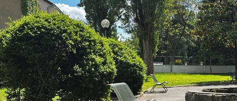 A paved area with two metal benches, a tree, and a hedge.