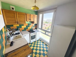 Une cuisine compacte avec des meubles en bois, une cuisinière, un évier et une fenêtre donnant sur des immeubles.
