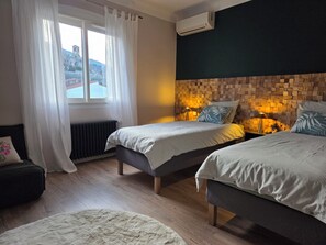 Une chambre avec deux lits simples, un radiateur, une fenêtre avec des rideaux, un appareil de climatisation et un mur orné d’un panneau en bois.