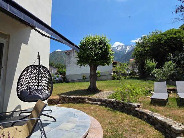 Une terrasse avec une table ronde, des chaises, un fauteuil suspendu et un mur en pierre.