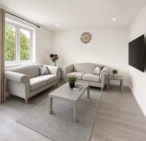 Uma sala de estar moderna com dois sofás, uma mesa de centro, uma mesa lateral, uma TV fixada na parede e uma luminária decorativa em formato de sol.