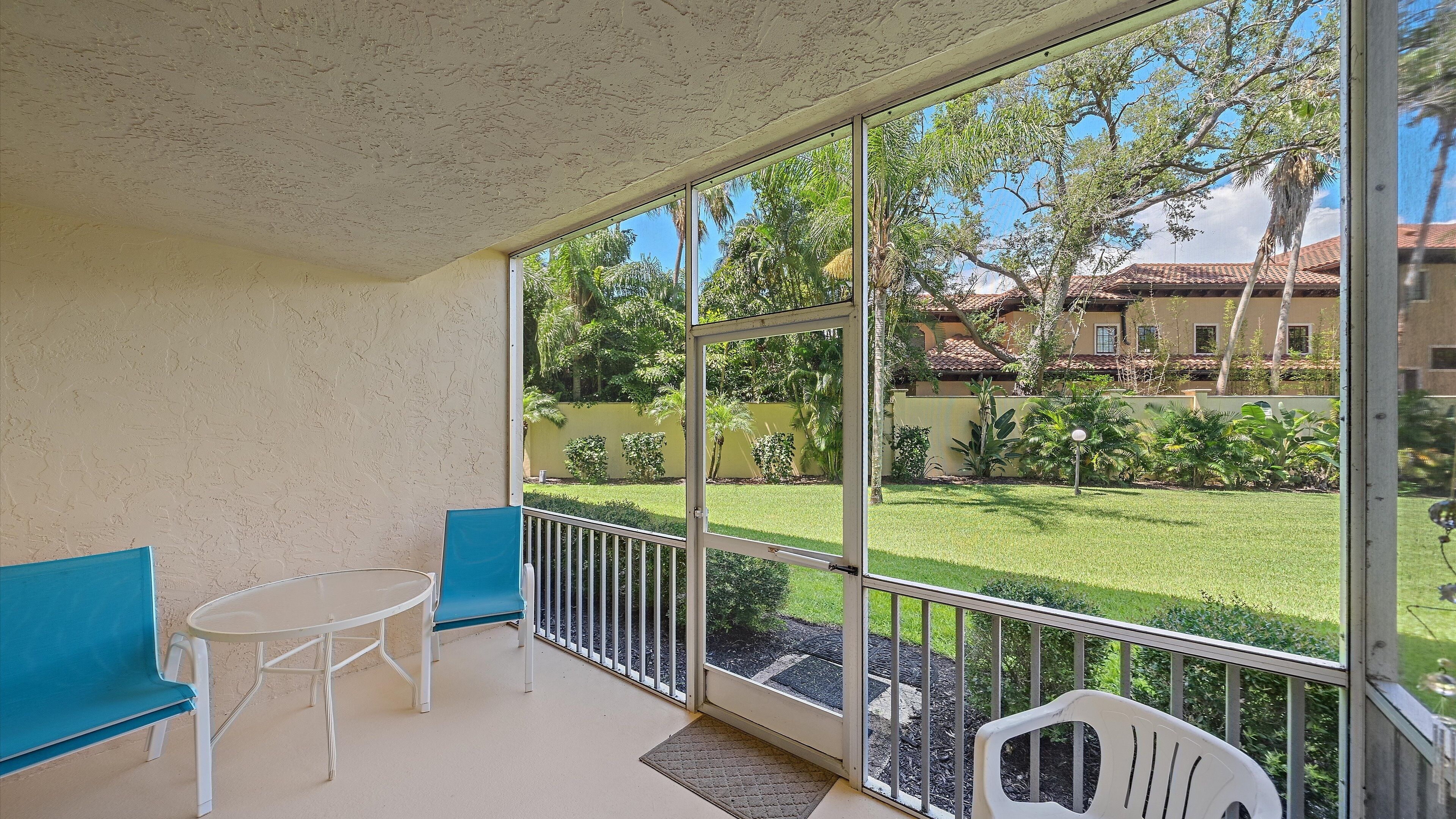 A balcony with a glass door, a small table, two chairs, and a view of a well-maintained garden and buildings in the background.