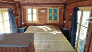 Ein Holzhüttenraum mit einem Bett, zwei Fenstern mit Vorhängen, einem Schreibtisch und einem Stuhl.