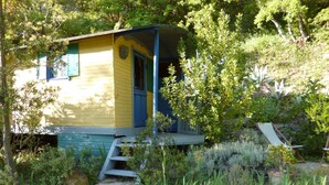 Eine kleine, eingeschossige Hütte mit Veranda, umgeben von üppigem Grün und einem Kiesweg.