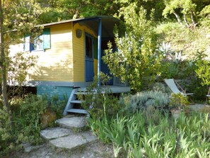 A small, single-story cabin with a porch, surrounded by lush greenery and a stone pathway. - Veyronne caravan (Aubres)