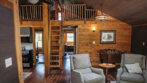 A wooden cabin interior with a loft bedroom, a dining area, and a kitchen.