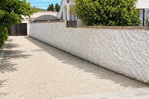 A gravel driveway leading to a gated entrance, a white textured wall, and a green bush. - Spacious apartment with terrace near the beach (Bizerte)