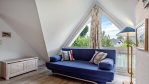 Una habitación con un sofá azul, un aparador de madera, una lámpara de pie y un amplio ventanal con vista a zonas verdes.