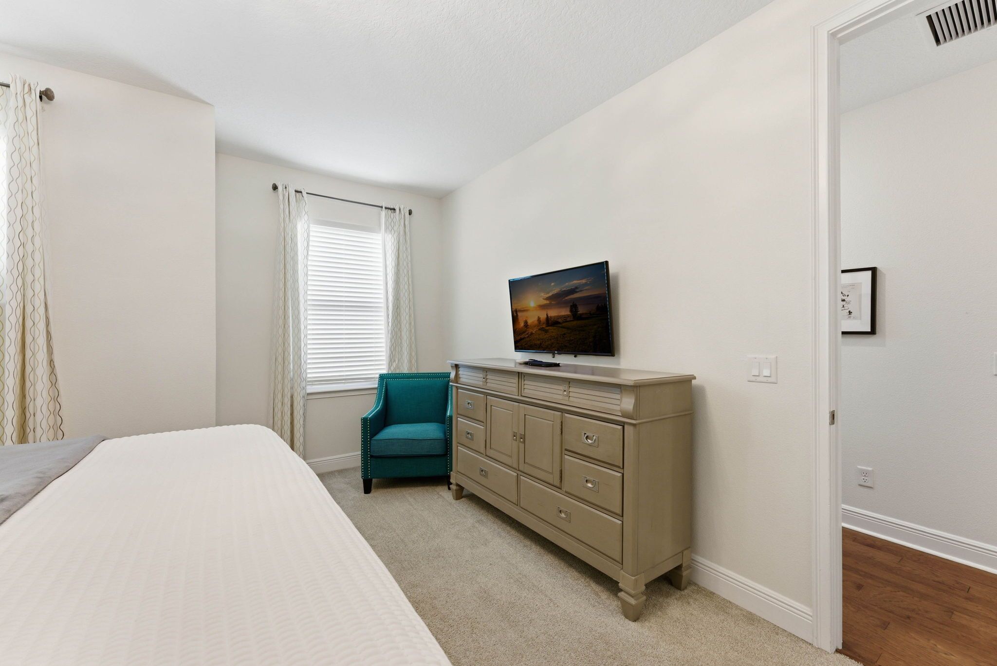 Un dormitorio con cama, cómoda, silla, ventana con cortinas y un cuadro en la pared.