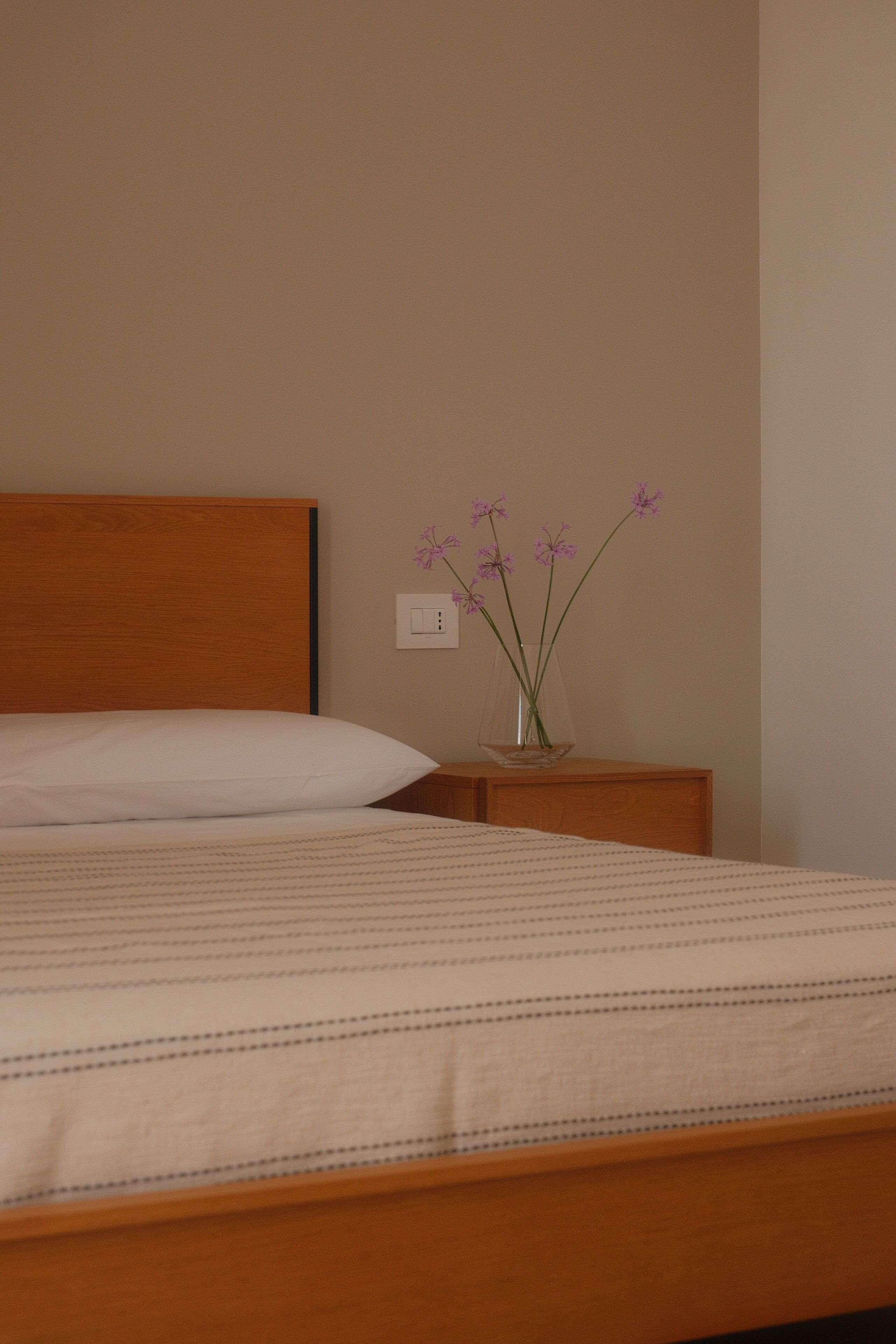 A bed with a wooden headboard, a bedside table with a vase of flowers, and a wall switch.