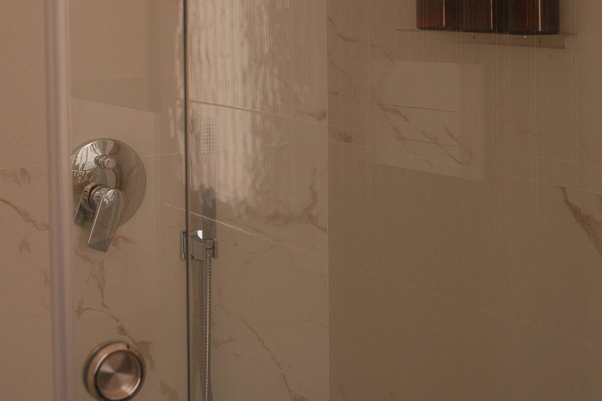 Ducha con cabezal de ducha manual, puerta de vidrio y repisa con tres botellas de champú, acondicionador y gel de baño.