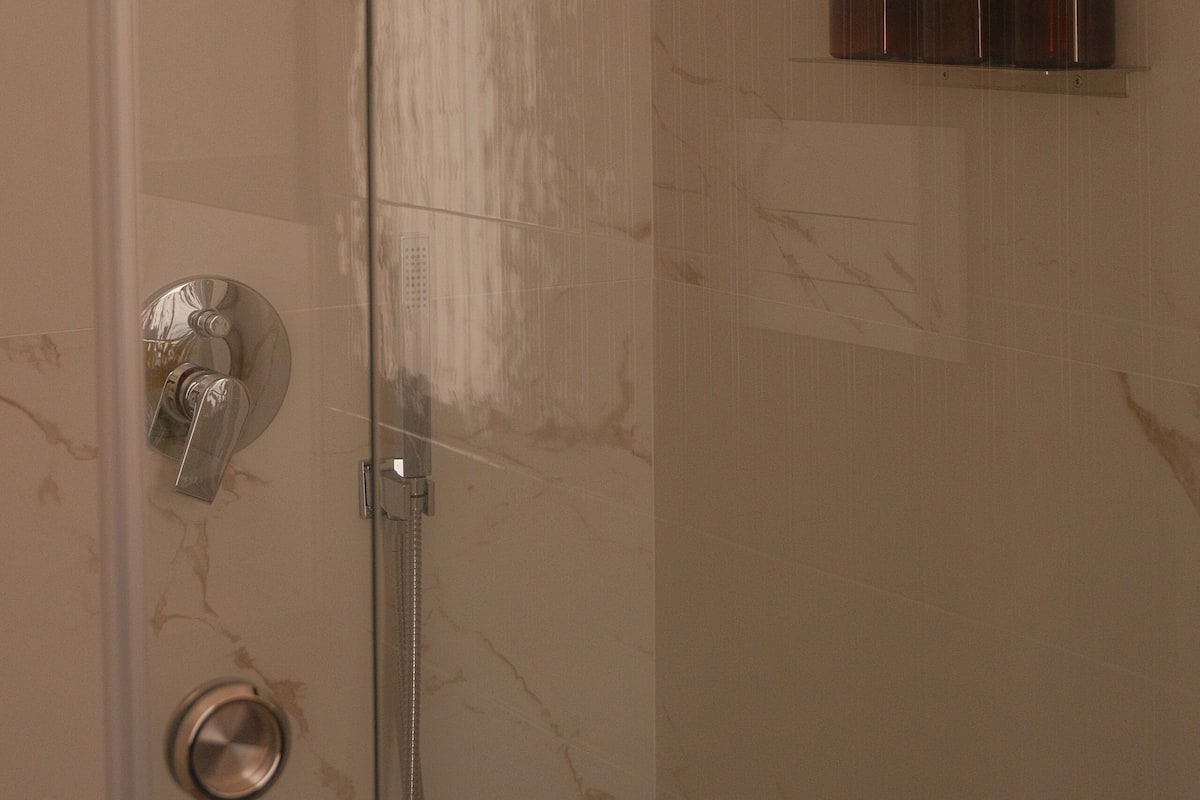 Ducha con cabezal de ducha manual, puerta de vidrio y repisa con tres botellas de champú, acondicionador y gel de baño.