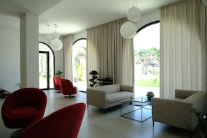 A modern living room with a sofa, red chairs, a coffee table, and large windows with curtains.