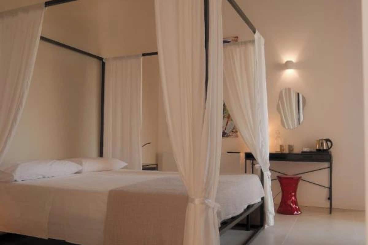 Una cama con dosel, cortinas blancas, una mesita de noche, un florero rojo y una lámpara de pared.