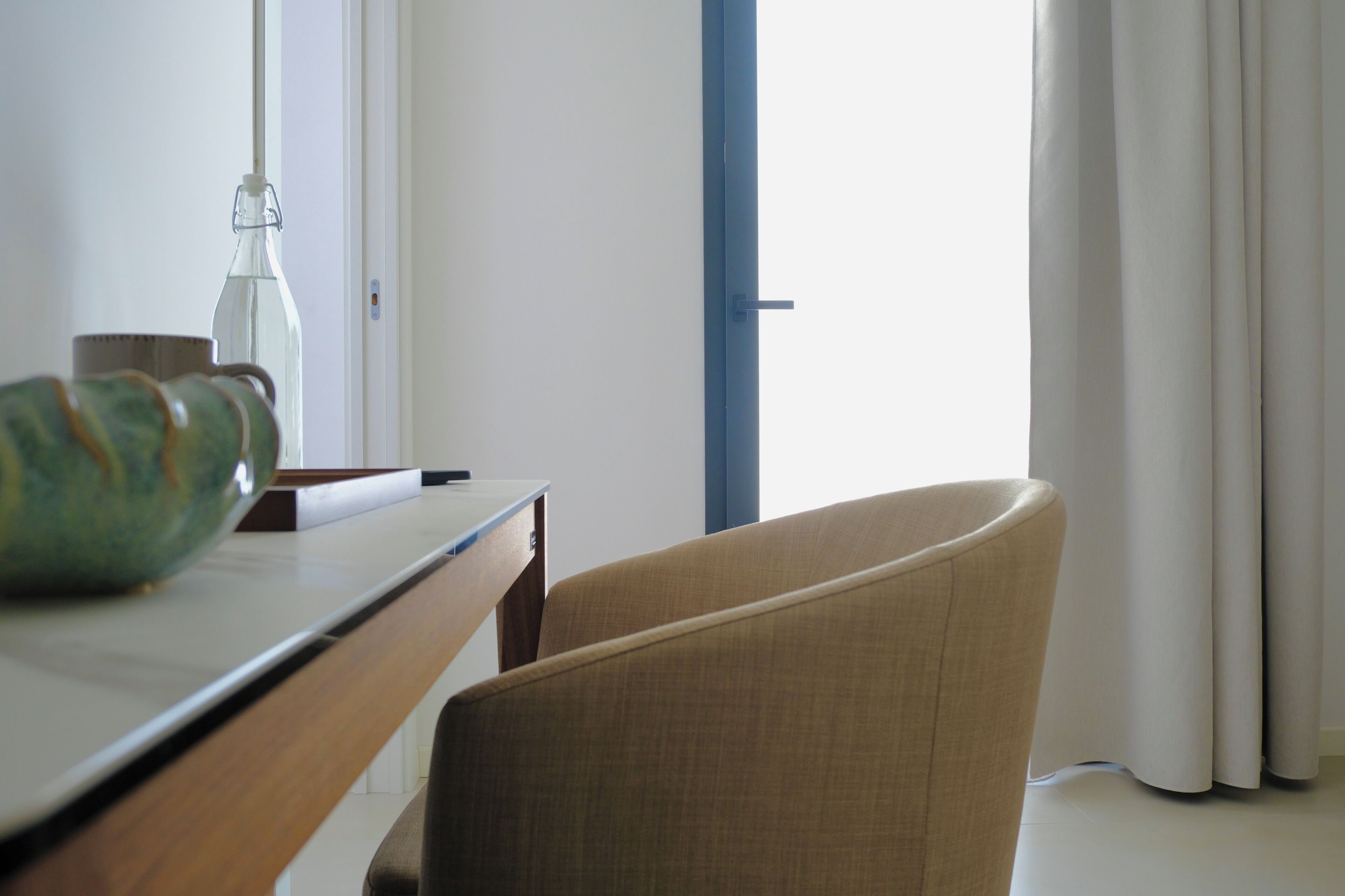 A room with a wooden desk, a chair, a glass bottle, and a window with curtains.