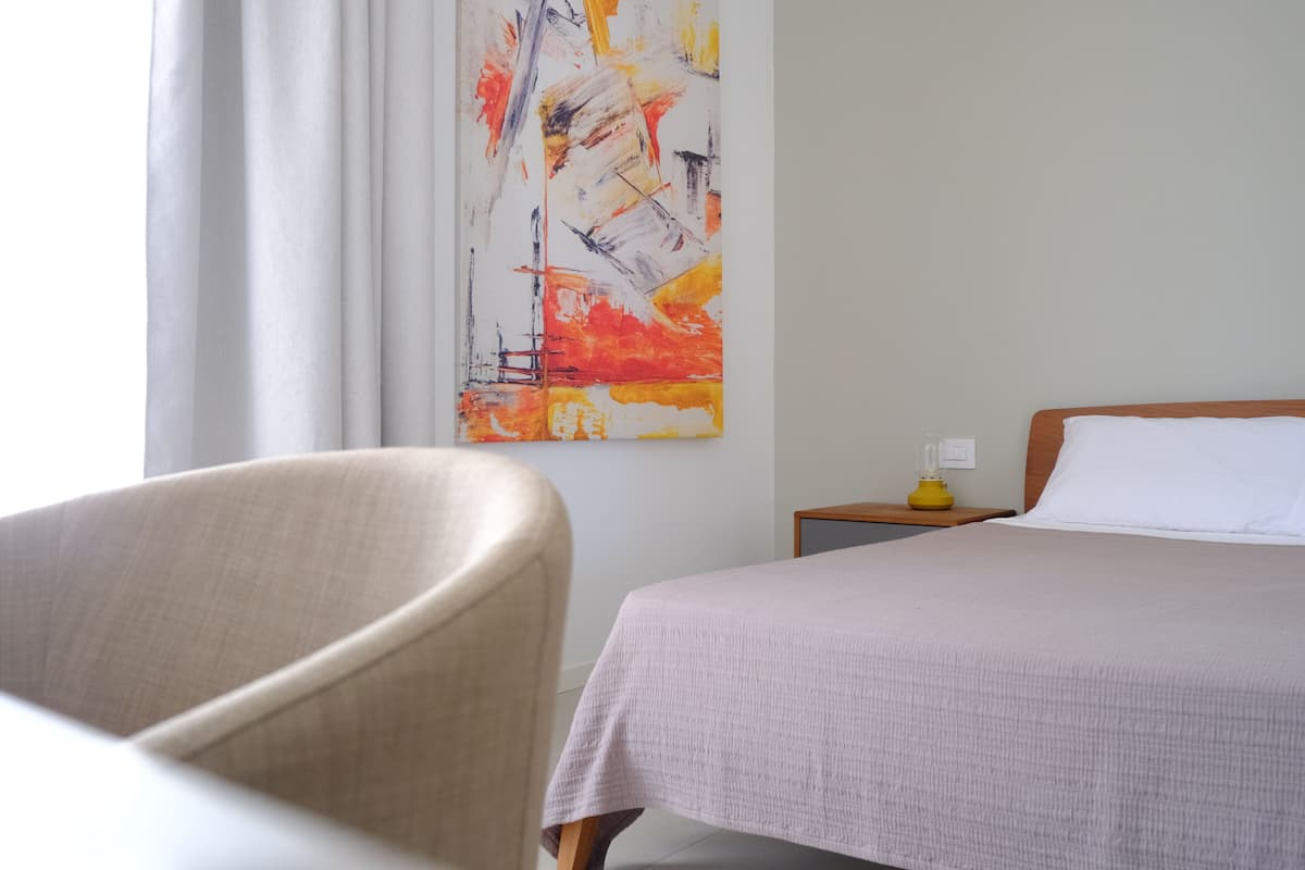 Habitación de hotel con cama, una silla, una mesita de noche y un cuadro en la pared.