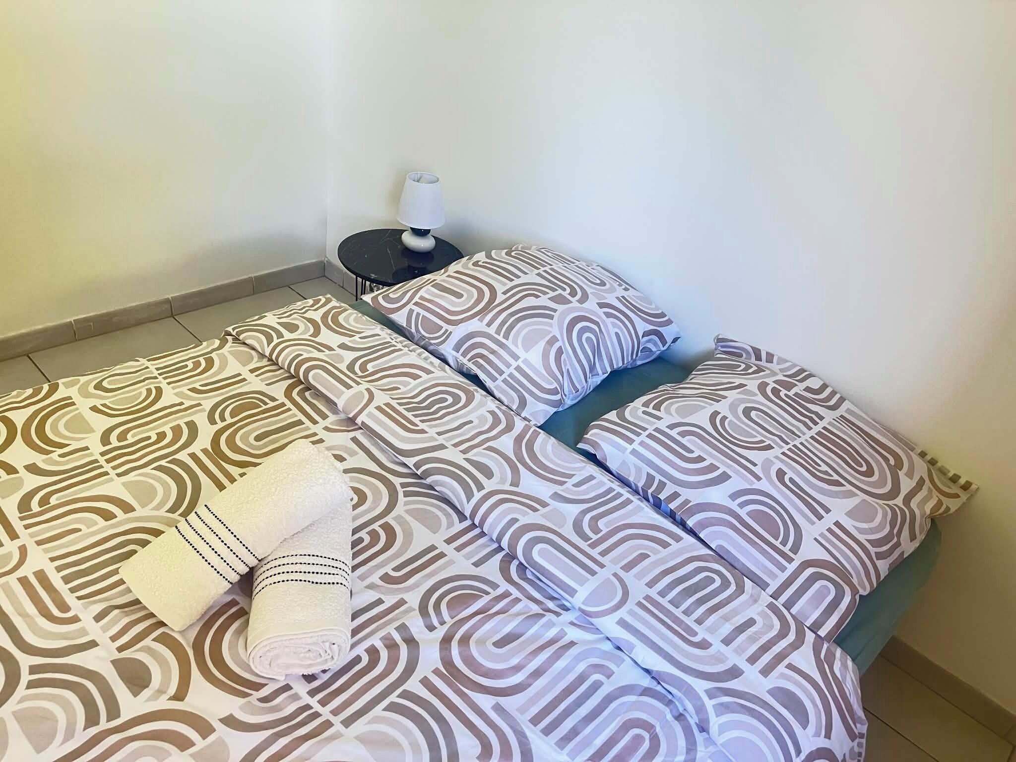 A neatly made bed with patterned bedding, a bedside table with a lamp, and a rolled towel.