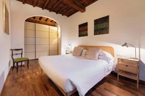 Une chambre avec un plancher en bois, un lit avec une literie blanche, une table de chevet avec une lampe et une chaise.